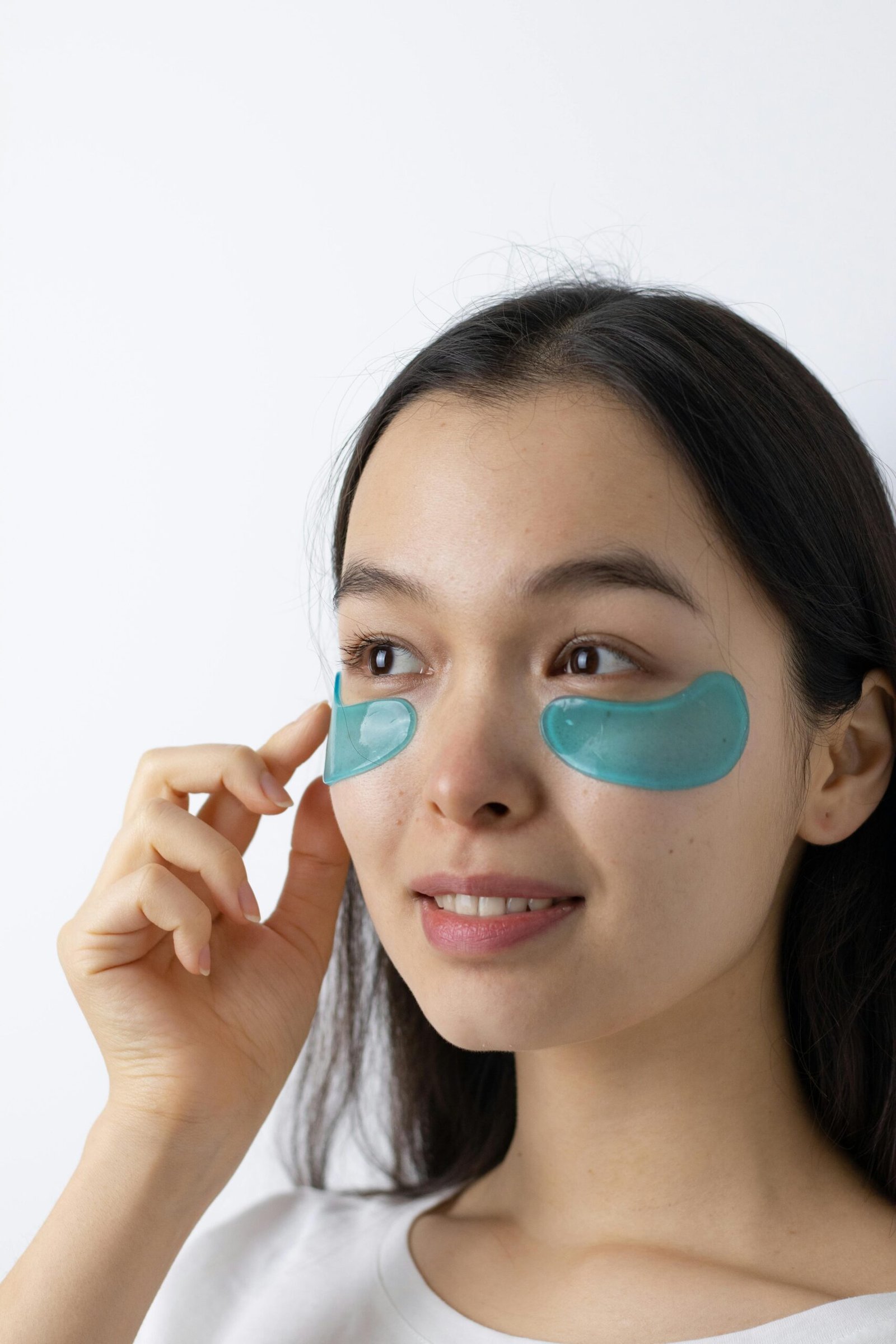 A young Asian woman applying turquoise under-eye masks for skincare.