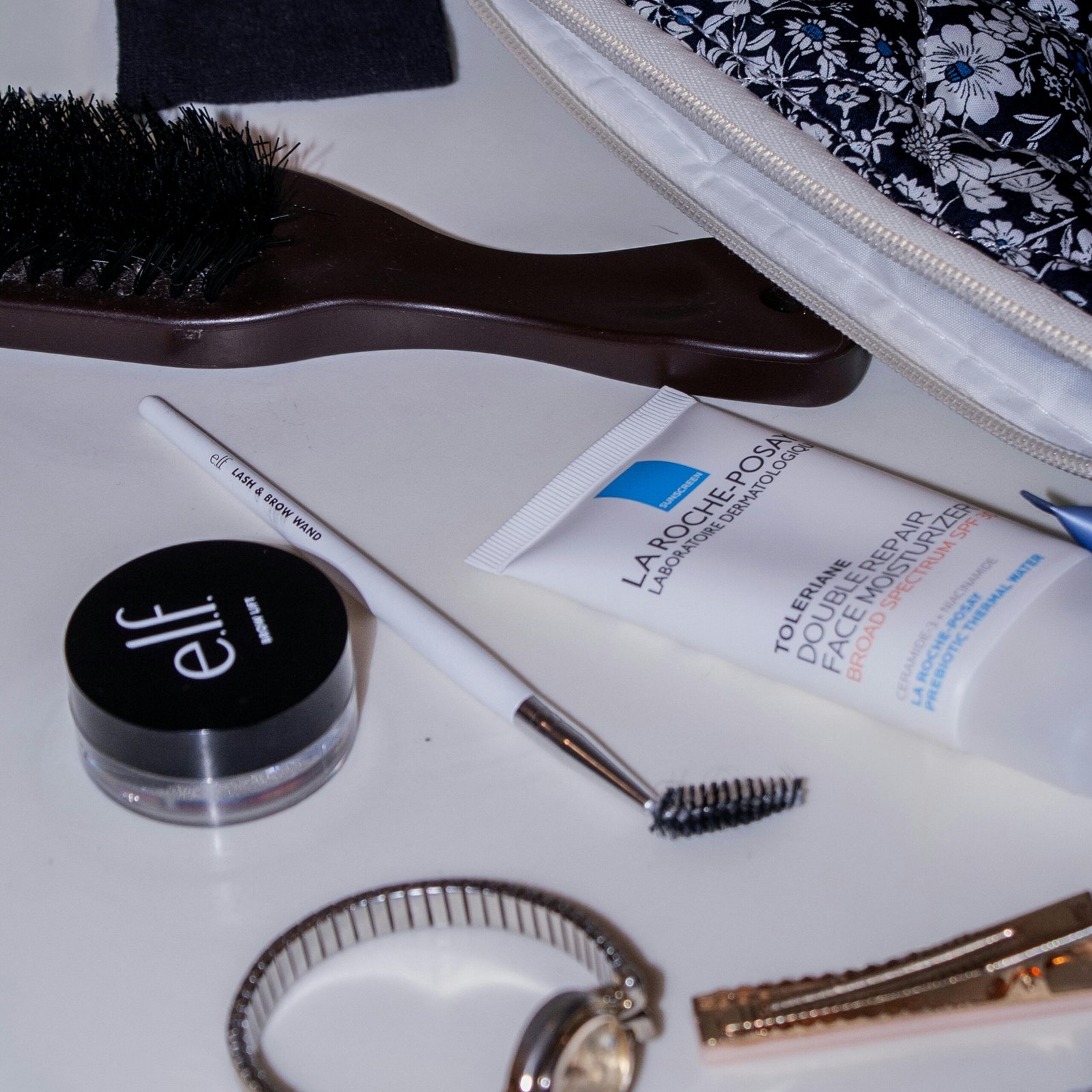 Flat lay of beauty essentials with a floral cosmetic bag, brush, watch, and makeup products.
