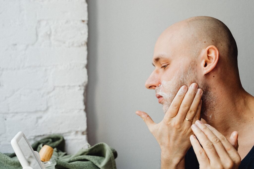 A bald man applies shaving cream to his face, focusing on personal grooming indoors.